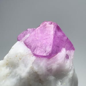 May include: Close-up of a vibrant pink crystal formation atop a white, textured mineral base. The crystal has a geometric shape with visible facets. The contrast between the pink and white creates a striking visual.