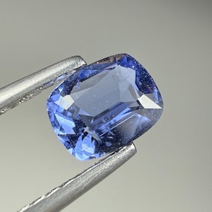 May include: A cushion-cut blue sapphire gemstone with a faceted surface, displaying a translucent medium-blue hue. The gemstone is held by silver-toned metal tweezers, showcasing its clear internal structure and polished edges in a bright light.