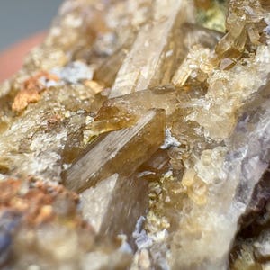 May include: Close-up of a mineral specimen featuring translucent, elongated crystals in shades of tan and brown. The crystals are clustered together, with a rough, textured matrix. The overall appearance suggests a natural, geological formation.