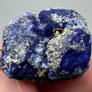 May include: A close-up of a raw, natural lapis lazuli gemstone. The stone is a deep, rich blue with patches of white and flecks of gold-colored pyrite. The surface is rough and textured, showcasing the natural beauty of the mineral.