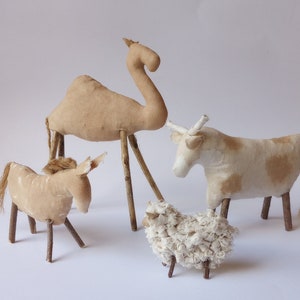 May include: A set of four handmade fabric animal figurines, including a camel, a cow, a donkey, and a sheep. The figurines are made from beige and white fabric and have twig legs.