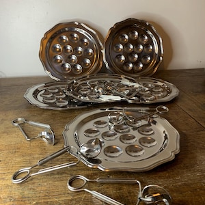 Peut inclure: Ensemble de plats à escargots et pinces argentés. Les plats ont des bords festonnés et des cavités pour maintenir les escargots. Les pinces sont conçues pour saisir les escargots. L'ensemble est disposé sur une surface en bois.