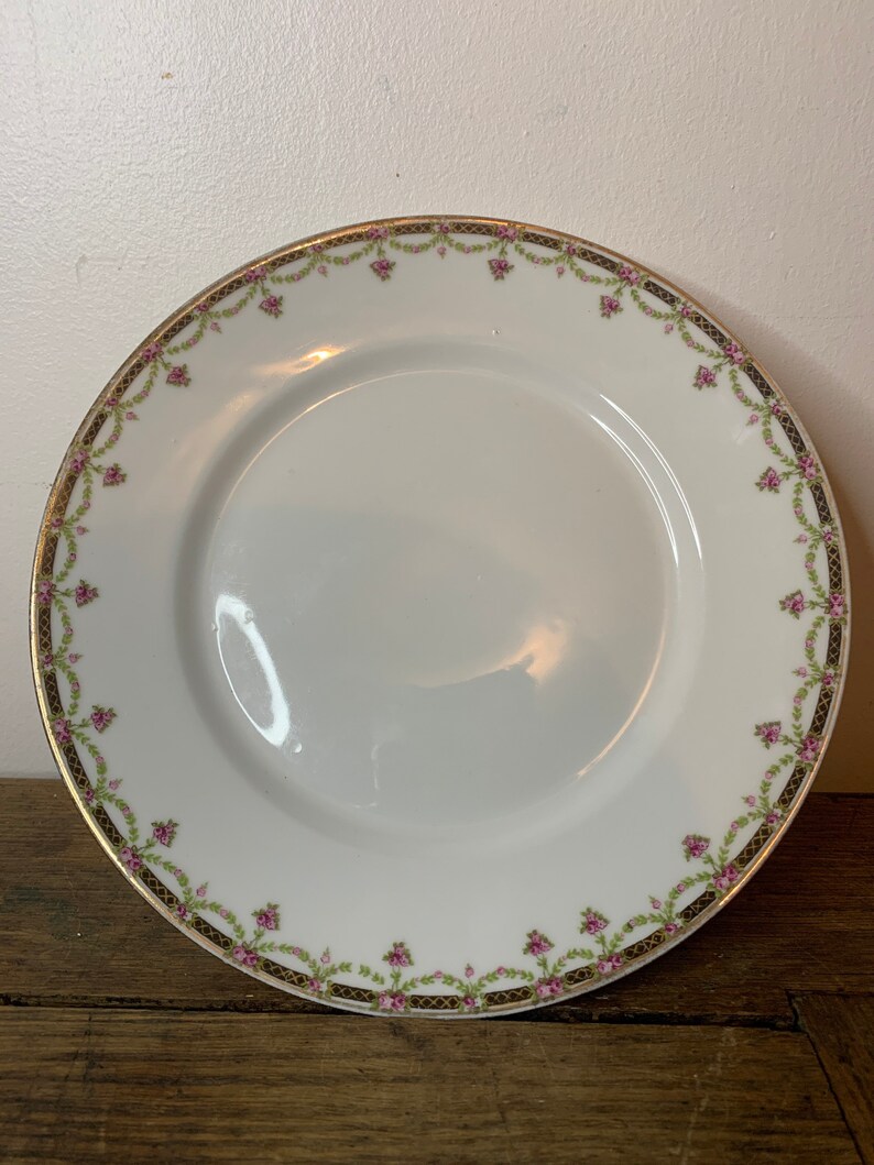 May include: White porcelain plate with a gold and black floral border. The border features pink flowers and green leaves.