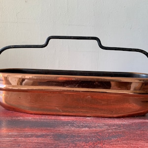May include: An oval copper planter with a black metal handle. The planter is made of polished copper and has a shiny finish.