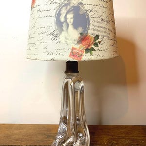 Może przedstawiać: Przezroczysta szklana lampa z unikalną, zakrzywioną podstawą i materiałowym abażurem. Abażur ma wzór vintage z portretem, różami i skryptem. Lampa to dekoracyjne oświetlenie.