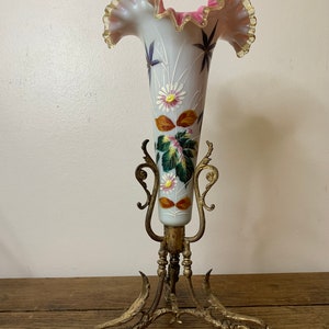 Może przedstawiać: Wazon z opalowego szkła z falbanką, ręcznie malowany we wzory kwiatowe, na mosiężnym stojaku. Wazon ma jasnoniebieski korpus z różowymi i złotymi detalami. Stojak ma około 30 cm wysokości.