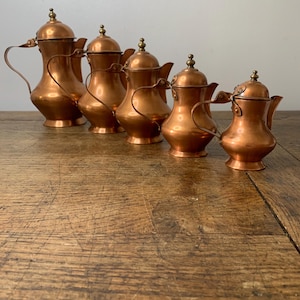 Peut inclure: Six théières en cuivre avec des poignées incurvées et des couvercles bombés sont disposées sur une surface en bois. Chaque théière est ornée d'un petit bouton rond sur le dessus. Le cuivre poli reflète la lumière.