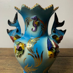 Könnte beinhalten: Eine blaue Keramikvase mit goldfarbenem Rand und goldenen Akzenten. Die Vase ist mit Fischen und einem Seepferdchen verziert. Die Vase ist etwa 20 cm hoch.