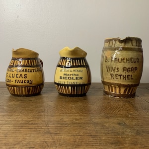 May include: Three vintage ceramic jugs with brown and yellow stripes. The jugs are decorated with text, including "MERIE-CHARCUTERIE", "GLUCAS", "LERS-FAUCON", "ARTICLES de MENAGE", "Marthe SIEGLER", "VIEUX-THANN Tel 576", "B. FAUCHEUX", "VINS AGAP", and "RETHEL".