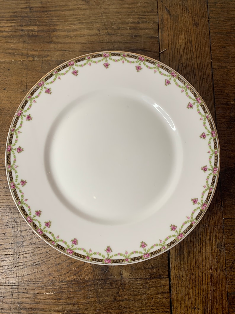 May include: A white porcelain plate with a gold and green floral border. The plate features a delicate floral design with pink flowers and green leaves.