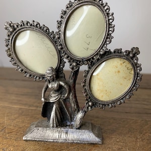 Peut inclure: Un porte-photos à trois cadres de couleur argentée avec un design orné. Les cadres ovales sont disposés en forme d'arbre, soutenus par une figurine sculptée. La base est rectangulaire, et le style général est vintage.