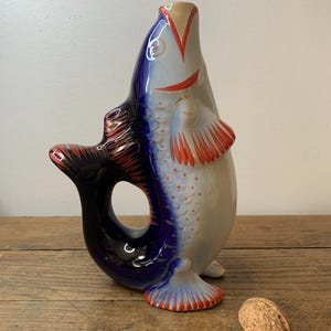 Op de afbeelding: Een keramische kan in de vorm van een vis, met blauwe en rode details op een witte basis. De kan heeft een grote opening en staat op een houten oppervlak, wat het unieke ontwerp en de kleuren benadrukt. Een decoratief item.