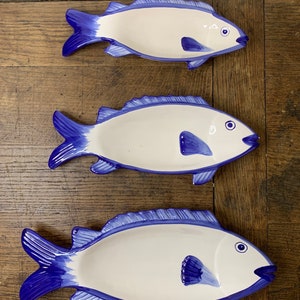 Puede incluir: Tres platos de servir de cerámica en forma de pez azul y blanco. Los platos están dispuestos en fila del más grande al más pequeño. Los peces tienen aletas y colas azules y cuerpos blancos con ojos azules.