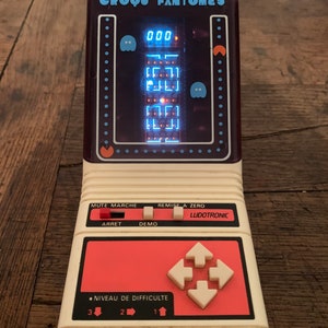 以下が含まれることがあります： 青いピクセル化されたゲームを表示する画面を備えた、ヴィンテージのポータブル電子ゲーム。 ゲームは白と赤で、「CROQU' FANTOMES」と「LUDOTRONIC」の文字が入っています。 方向ボタンとレベルインジケーターがあります。