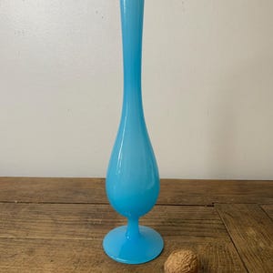 Puede incluir: Un jarrón de cristal azul claro, esbelto, con base acampanada y cuello largo. El jarrón mide unos 30 cm de alto y está sobre una superficie de madera. Una nuez marrón está junto al jarrón.