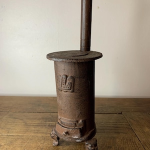 Può includere: Una piccola stufa antica in ghisa con corpo cilindrico e piano rotondo. La stufa ha un lungo camino verticale e poggia su tre piedi decorati. L'esterno è di colore marrone usurato, con un emblema decorativo sul davanti.