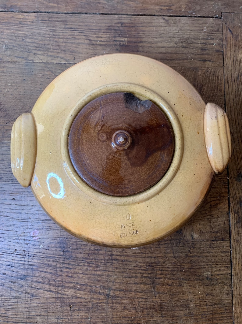 K&ouml;nnte beinhalten: Eine gelbe Keramik-Auflaufform mit einem braunen Deckel und zwei Griffen. Die Form hat eine wei&szlig;e Aufschrift mit der Aufschrift "Made in France".