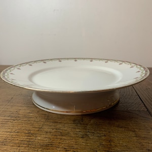 May include: White ceramic cake stand with a gold rim and a delicate floral design in shades of green and pink.