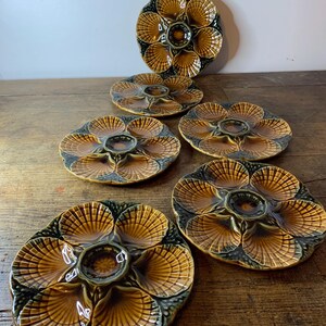 Op de afbeelding: Set van zes keramische oesterborden. Elk bord heeft een bruin en groen schelpmotief met individuele compartimenten. De borden zijn gerangschikt op een houten oppervlak, wat hun unieke vorm en kleur benadrukt.