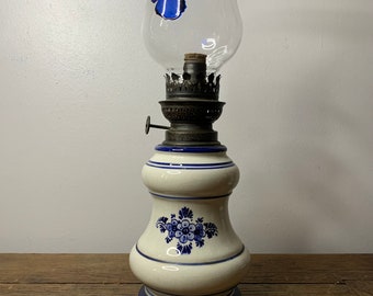 May include: A white ceramic oil lamp with blue and white floral designs. The lamp has a clear glass chimney with a blue butterfly decal and the word "Caussade" written on it.