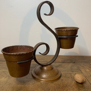 Peut inclure: Un bougeoir décoratif en métal de couleur cuivre avec deux petits pots. Le bougeoir a un design en volutes et une base ronde. Les pots ont une surface texturée et un bord décoratif. Une noix est posée sur la surface en bois.