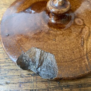 K&ouml;nnte beinhalten: Ein brauner Keramikdeckel mit einem Knopf oben. Der Deckel hat einen Abplatzer an der Kante.