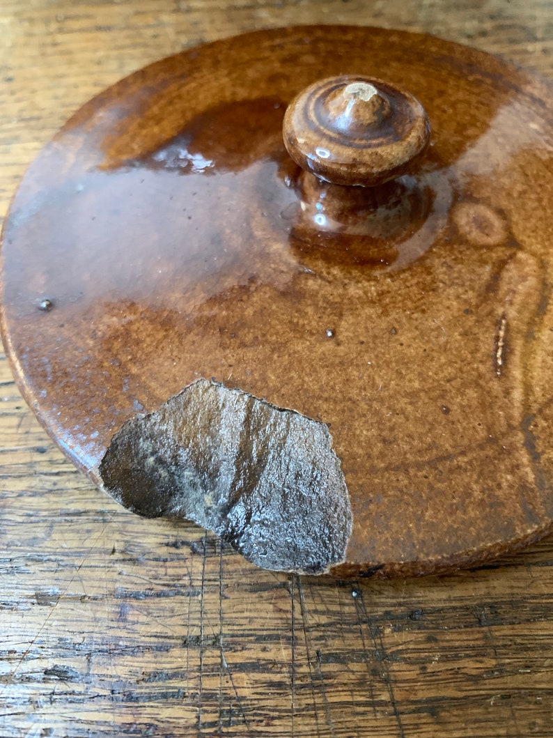 K&ouml;nnte beinhalten: Ein brauner Keramikdeckel mit einem Knopf oben. Der Deckel hat einen Abplatzer an der Kante.