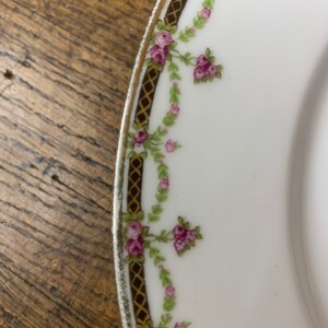 May include: A white porcelain plate with a gold rim and a delicate floral design. The design features pink roses and green leaves on a white background.