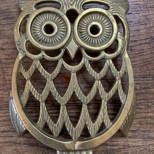 Peut inclure: Un dessous de plat en laiton en forme de hibou avec un design ajouré, des plumes gravées détaillées, de grands yeux circulaires à motifs radiaux et un bec pointu. La surface métallique présente une finition dorée avec une patine naturelle.