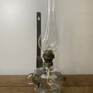 Könnte beinhalten: Eine antike Öllampe aus Glas mit einer Wandhalterung aus Metall. Die Lampe hat einen klaren Glasschirm und einen Glasbehälter. Die Lampe steht auf einer Holzoberfläche mit einer Walnuss.