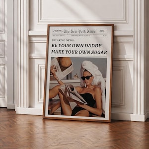 Puede incluir: Impresión enmarcada con un titular de periódico: "BE YOUR OWN DADDY MAKE YOUR OWN SUGAR". La imagen muestra a una persona con gafas de sol y una toalla, leyendo un periódico. El marco es de color marrón cálido.