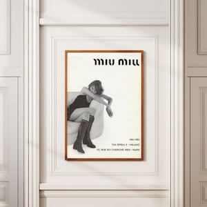 Peut inclure: Une affiche murale rectangulaire dans un cadre en bois présentant une photographie en noir et blanc d'une personne assise sur un canapé, vêtue d'une robe sombre et de bottes hautes. Le texte MIU MIU figure en haut, avec MIU MIU, VIA SPIGA 5, MILANO et 10, RUE DU CHERCHE MIDI, PARIS en bas.