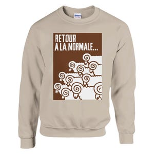 May include: A beige crewneck sweatshirt featuring a brown rectangular graphic on the chest. The graphic displays white stylised sheep with curled horns and the text "RETOUR A LA NORMALE..." printed in white capital letters above the sheep.