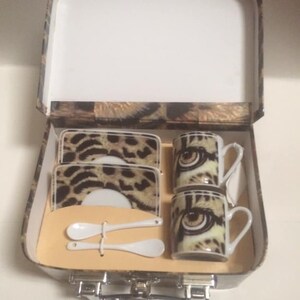 May include: A leopard print tea set in a suitcase-style box. The set includes two mugs, two saucers, two small spoons, and two coasters. The mugs and coasters feature a close-up of a leopard's eye.