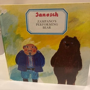 Può includere: Copertina di un libro per bambini con il titolo "Zampano's Performing Bear" di Janosch. L'illustrazione mostra un uomo con un cappotto blu e un orso bruno su sfondo giallo e bianco.
