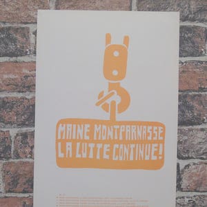 Puede incluir: Póster naranja y blanco con un gráfico estilizado de grúa. El texto "MAINE MONTPARNASSE LA LUTTE CONTINUE!" está en letras bloque naranjas. El póster está contra una pared de ladrillos.