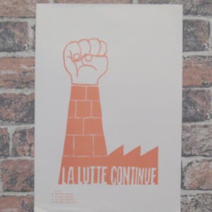 Puede incluir: Póster naranja y blanco con un puño en alto, un brazo con forma de ladrillos y la frase francesa «LA LUTTE CONTINUE». El diseño incluye una silueta de fábrica y texto en la parte inferior. El póster se muestra contra una pared de ladrillos.