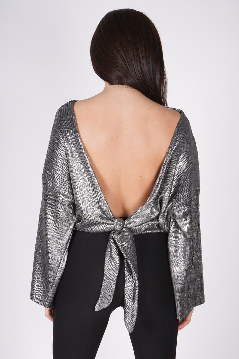 May include: A silver metallic top with long sleeves and a low back. The top is tied in a knot at the back.