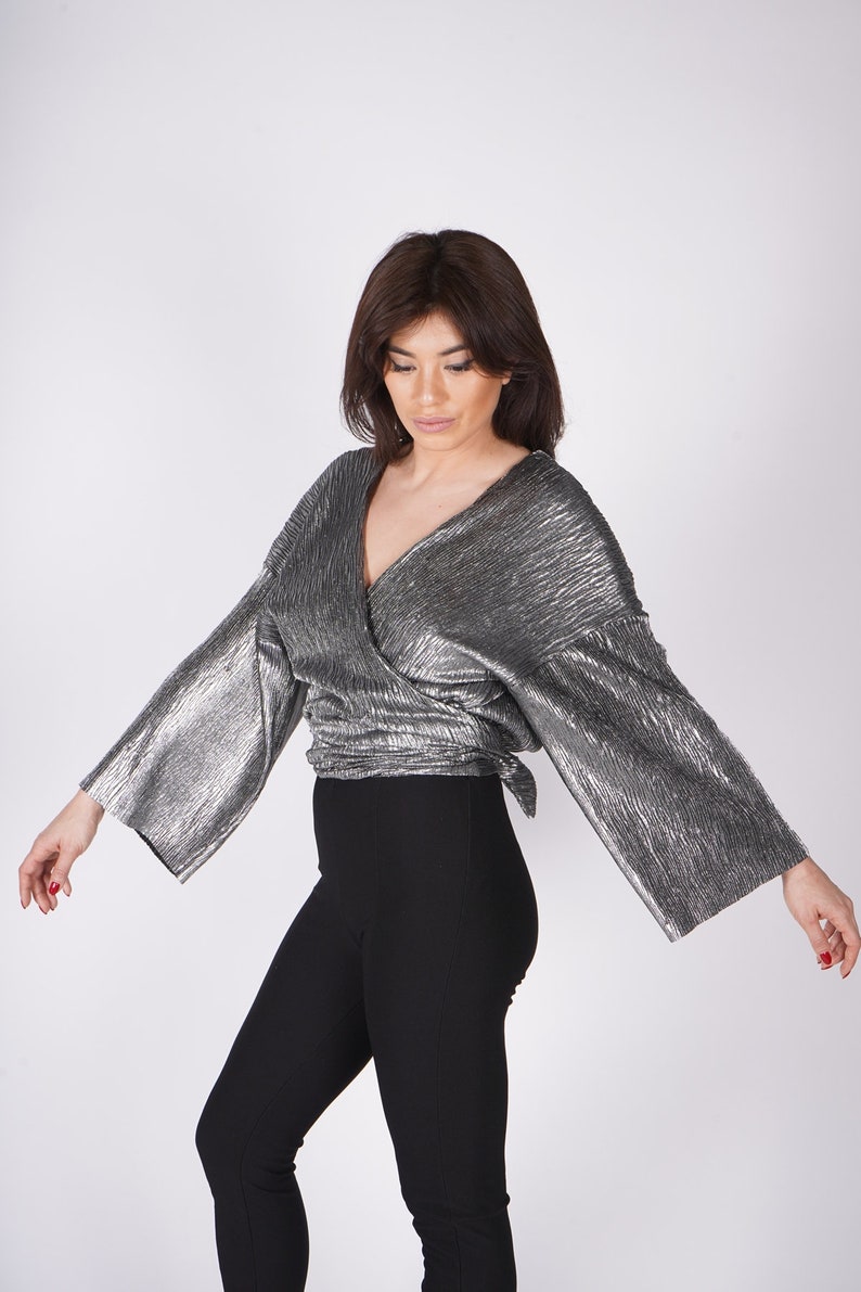 May include: A silver metallic wrap top with long sleeves and a V-neckline. The top is tied at the waist and has a flowing, relaxed fit.