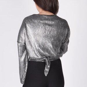 May include: A silver metallic top with long sleeves and a tie at the back. The top is worn with black leggings.