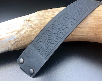 Op de afbeelding: Zwart leren armband met een gedetailleerd reliëfpatroon van geometrische vormen en spiralen. De armband heeft twee zilveren metalen klinknagels.