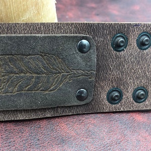 Op de afbeelding: Een bruin leren armband met een donkerbruin leren patch met een gegraveerd verenpatroon. De armband is vastgemaakt met metalen klinknagels.