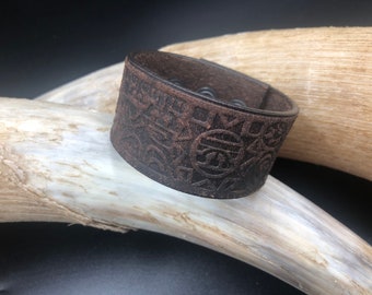 Op de afbeelding: Een bruin leren armband met een gegraveerd geometrisch patroon op het oppervlak. De armband is gemaakt van echt leer en heeft een rustieke, vintage uitstraling.