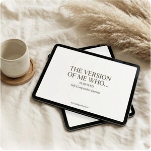 May include: Two black tablets with white screens displaying text: "THE VERSION OF ME WHO... SURVIVED. Self-Compassion Journal." A ceramic mug and wooden coaster sit to the left. Dried ornamental grass is in the background. The journal is by healingwithroseco.