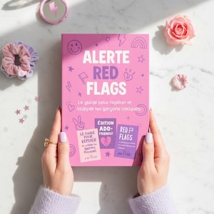 May include: A pink book titled "ALERTE RED FLAGS" with illustrations of a smiley face, lightning bolt, and rainbow. The book is held by a person wearing a lavender sweater. A pink compact mirror, scrunchies, and a rose are also visible.