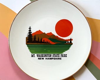 May include: A white ceramic plate with a gold rim featuring a landscape design. The design includes a red sun, mountains, trees, and the text "MT. WASHINGTON STATE PARK NEW HAMPSHIRE". The plate is on a colorful striped background.