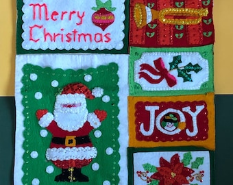 May include: A white felt Christmas stocking with a green, red, and yellow patchwork design. The stocking features embroidered images of Santa Claus, a Christmas ornament, a trumpet, a holly sprig, a snowman, and a poinsettia. The stocking also has the words "Merry Christmas" and "Joy" embroidered on it.