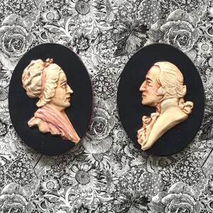 May include: Two oval-shaped cameo portraits. One features a woman in profile with a pink scarf and a white wig. The other depicts a man in profile with a white wig and a cream-colored jacket. Both are set against a black background.