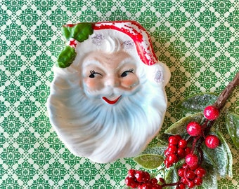 May include: A white ceramic Santa Claus dish with a red hat and a white beard. The dish is on a green and white patterned background.