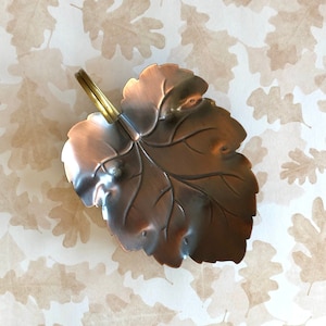 May include: A copper leaf-shaped dish with a brass handle. The dish is designed to look like a single leaf with veins and a pointed tip.